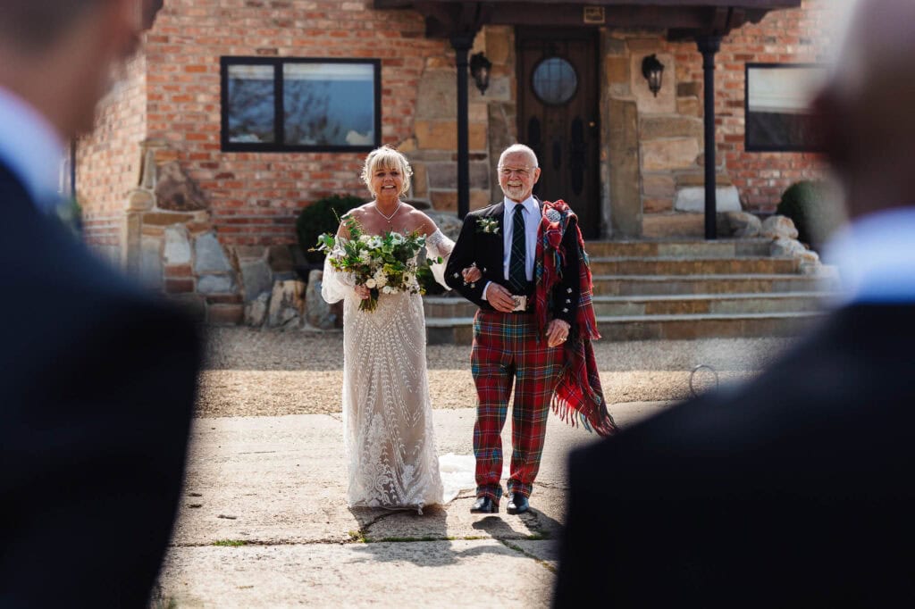 Elegant bride walking down the aisle with her father at a wedding ceremony outside BGS Weddings, showcasing beautiful wedding portraits and Scottish wedding traditions in a picturesque outdoor setting.