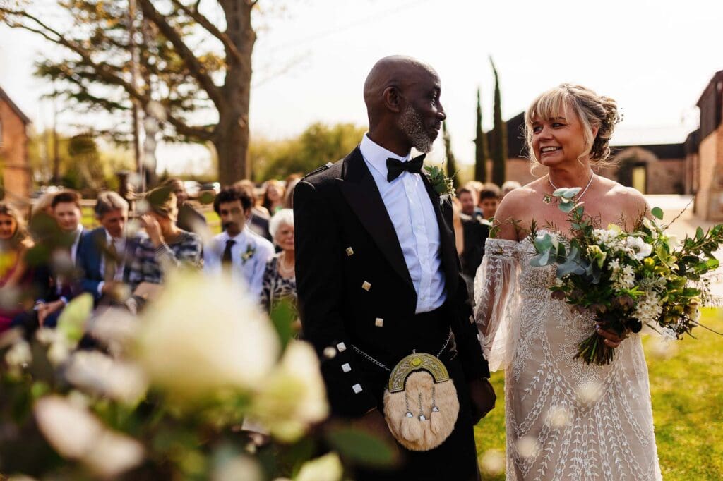 Elegant wedding ceremony with bride and groom exchanging vows outdoors, surrounded by guests, lush greenery, and natural light, captured by BGS Weddings.