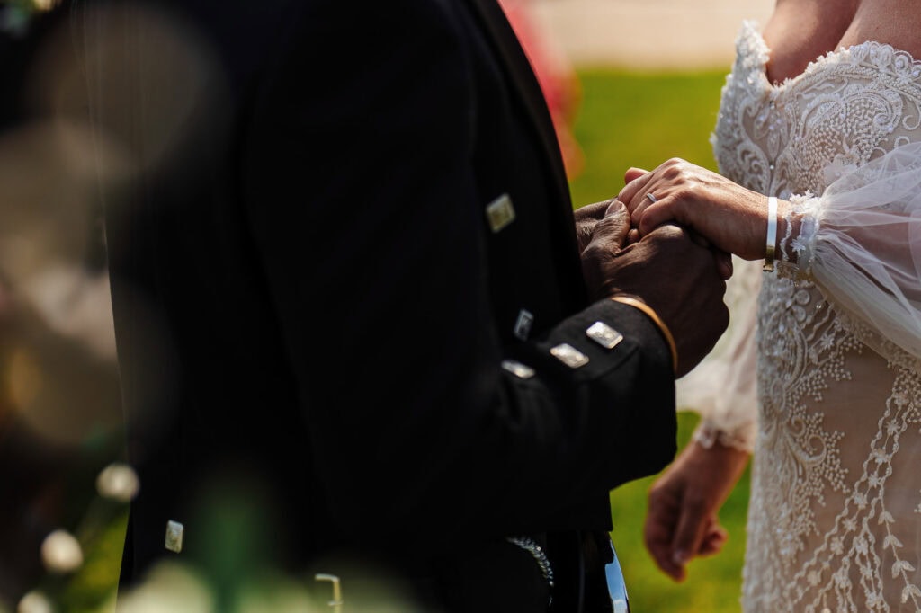 Delicate wedding ring exchange between a bride in an elegant lace gown and a groom in a black tuxedo outdoors at BGS Weddings. Stunning wedding ceremony captured beautifully.