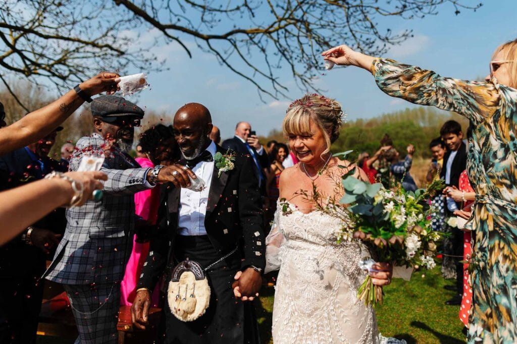 Colourful outdoor wedding celebration with confetti, guests throwing flower petals, bride and groom smiling, in vibrant spring setting with scenic natural backdrop.