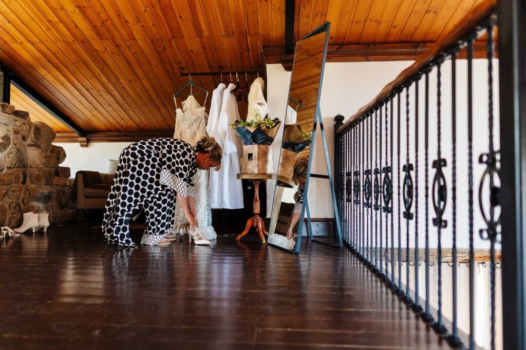Elegant bride preparing wedding shoes in rustic bridal suite with wooden ceiling and vintage decor, part of BGS Weddings services in the UK.