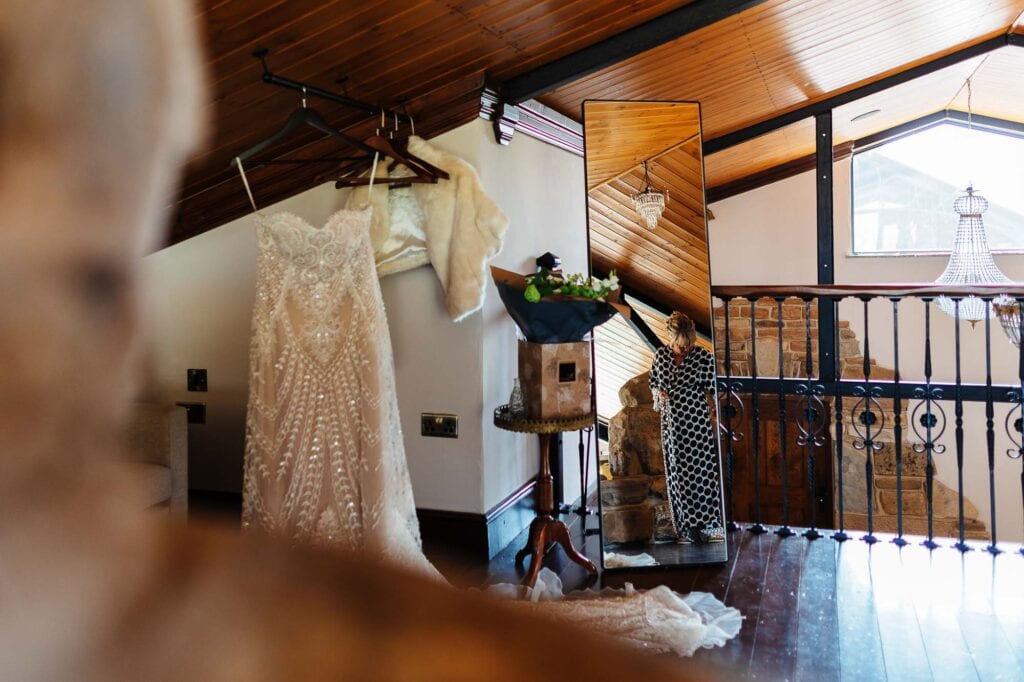 Elegant wedding dress hanging with fur stole, bridal accessories, and a bridesmaid preparing in a rustic wedding venue with wooden ceiling, stone staircase, and vintage decor.