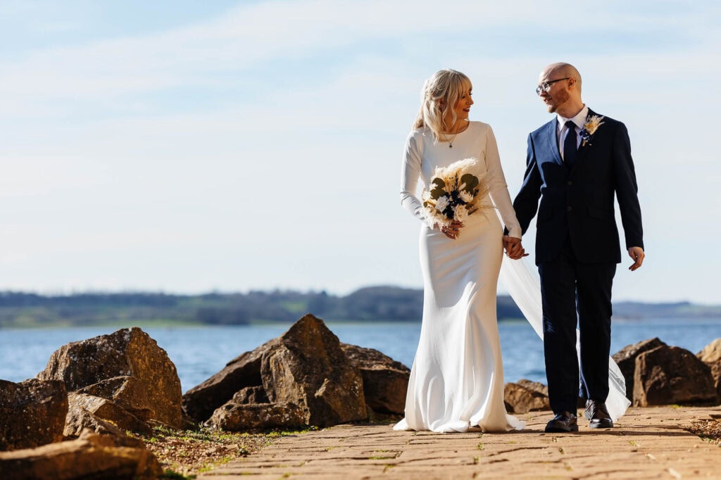 Elegant wedding couple walking hand in hand along the shoreline, dressed in classic wedding attire with a scenic water backdrop.