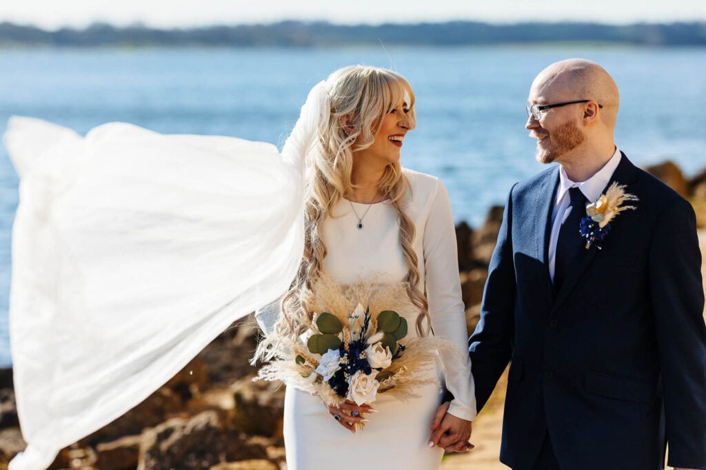Happy bride and groom holding hands by the seaside during their wedding ceremony, with scenic water background and rocky shoreline, capturing a joyful moment.