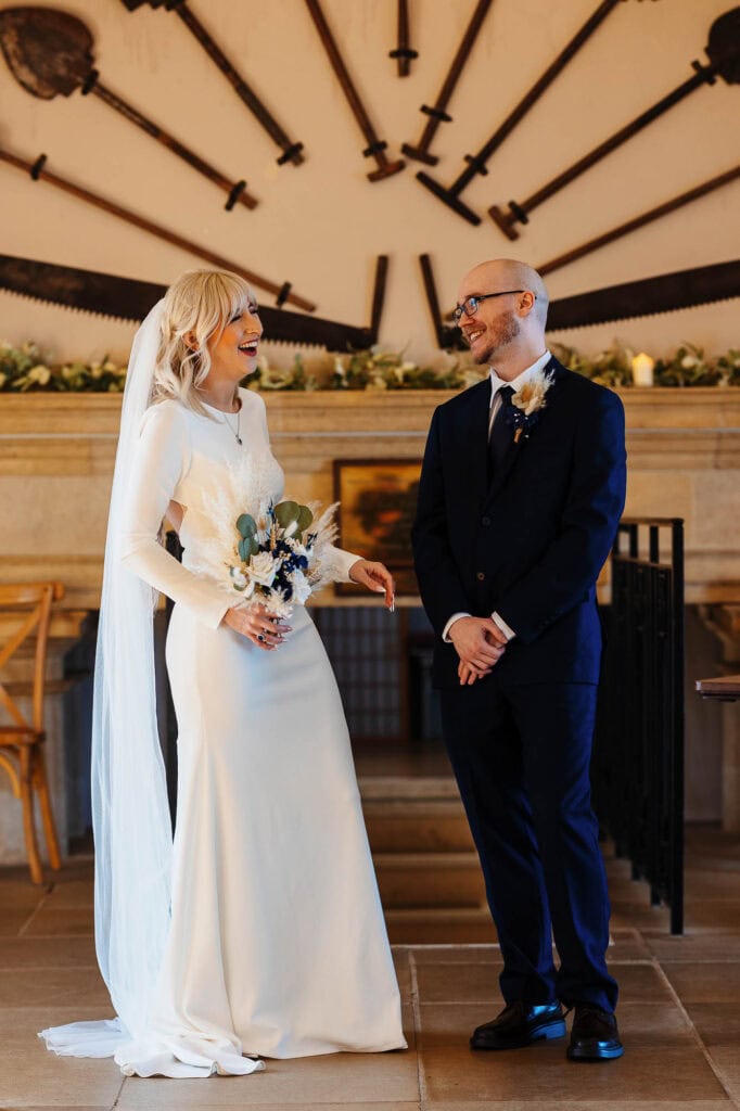 Bride and groom exchanging vows indoors with a large clock and vintage decor in the background. Beautiful moment captured during a wedding ceremony at BGS Weddings.