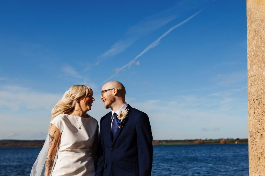 Happy bride and groom smiling at each other during their wedding ceremony outdoors, with a scenic lakeside background and clear blue sky.