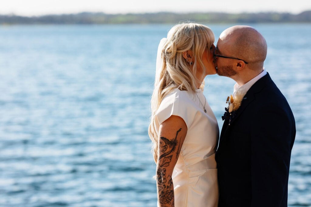 Romantic wedding couple sharing a kiss by the water, with scenic lake background, celebrating their special day in a beautiful outdoor setting.