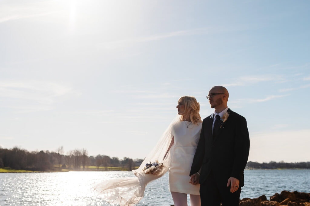 A bride and groom holding hands by a lakeside during sunset, capturing a romantic moment on their wedding day. The bride wears a white dress with a flowing veil, and the groom is dressed in a dark sui.
