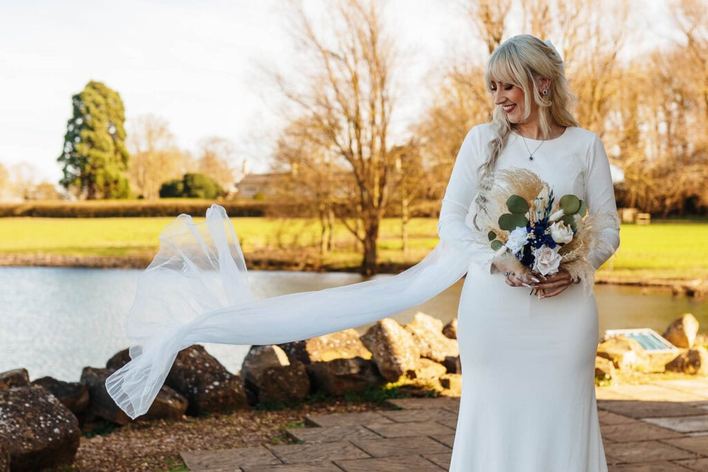 Bride with flowing veil and bouquet by the lake in a scenic outdoor setting.
