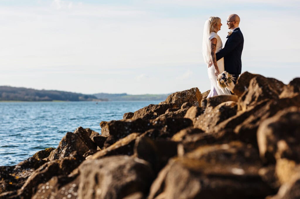 Romantic wedding couple standing on rocks by the sea, capturing a beautiful moment during their wedding ceremony with scenic water and coastline in the background.