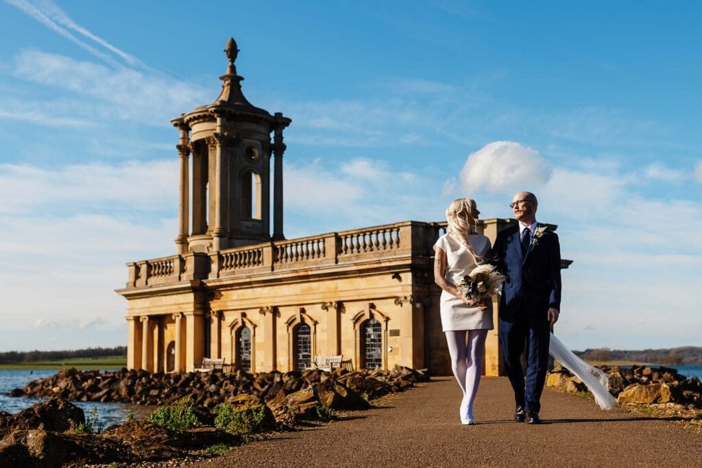 Beautiful seaside wedding ceremony at BGS Weddings with a historic building backdrop and a couple walking along the coast.