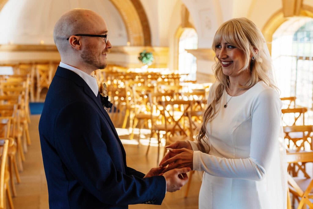 A couple exchanging vows during a wedding ceremony at BGS Weddings, showcasing a joyful moment in a beautifully lit, classic venue with wooden chairs and arched windows.