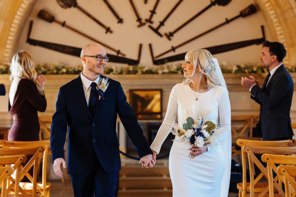 A couple holding hands during their wedding ceremony at BGS Weddings, with guests and a large clock in the background, showcasing a beautiful and joyful moment.