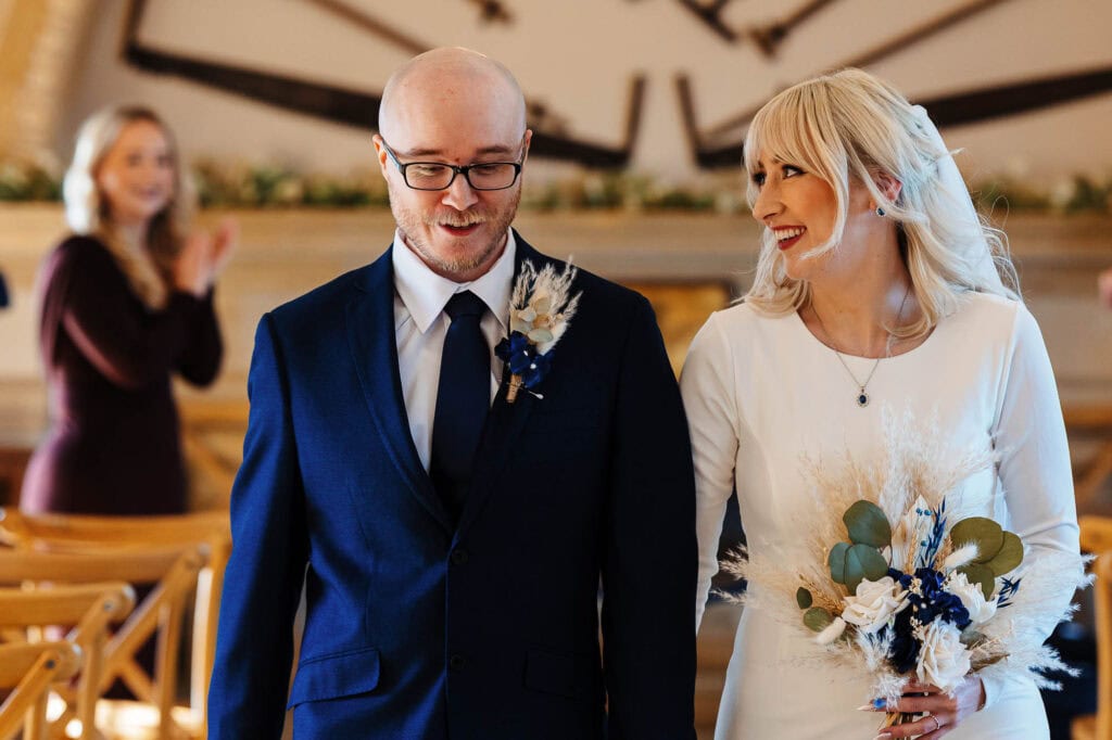 Happy bride and groom during their wedding ceremony at BGS Weddings, with a joyful guest in the background, capturing a special moment in a beautifully decorated venue.