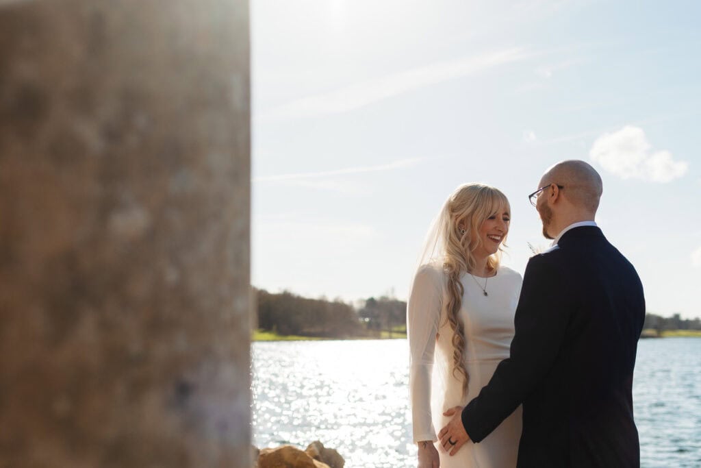 Happy bride and groom holding hands by the lake during their wedding, with sunlight reflecting on the water and a clear sky, capturing a romantic outdoor wedding moment.