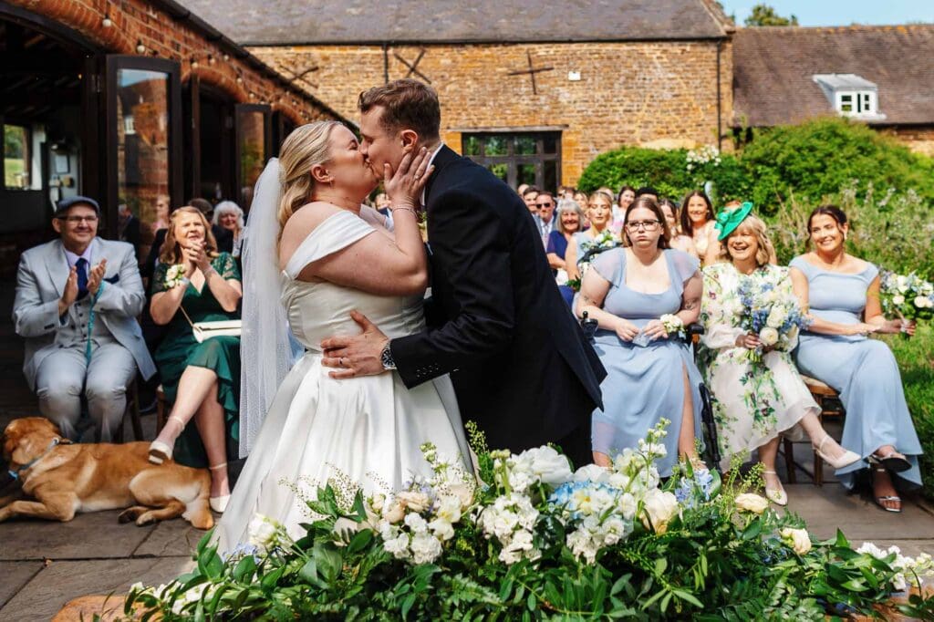 A1. Happy wedding couple sharing a kiss during their outdoor ceremony with friends and family, surrounded by floral decorations and a rustic brick building in the background.
