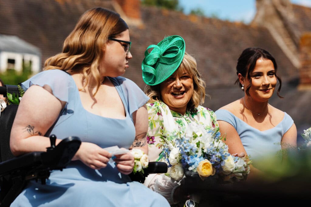 Elegant women dressed in vintage-style attire at a wedding ceremony outdoors, with a scenic background of rooftops and clear skies.