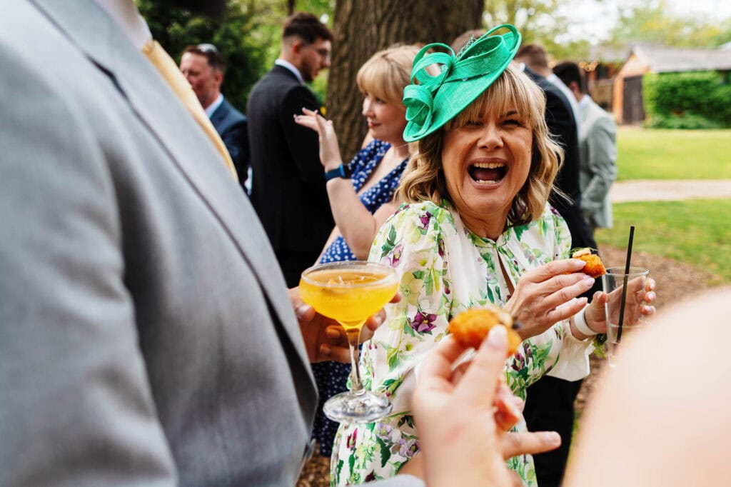 Joyful outdoor wedding reception with guests enjoying drinks and canapés, featuring a woman in a floral dress and teal fascinator smiling and engaging with friends in a lush garden setting.
