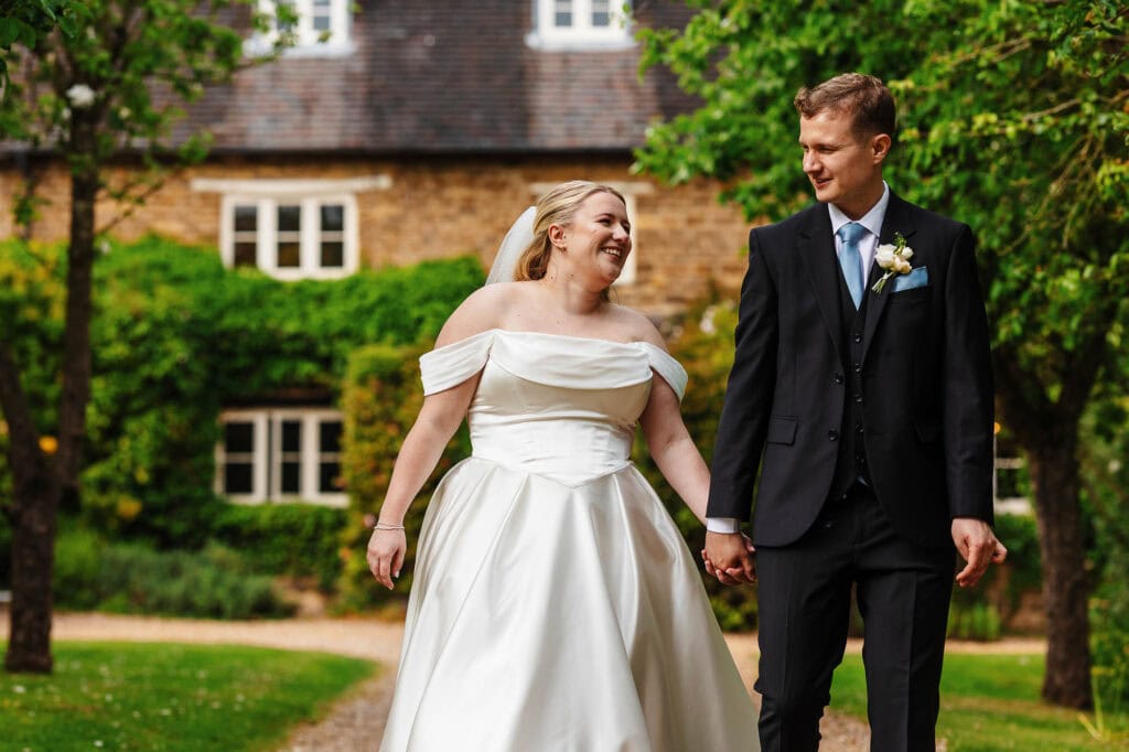 Happy bride and groom walking hand-in-hand outdoors in wedding attire, surrounded by lush greenery and historic brick buildings, celebrating their special day in a picturesque setting.