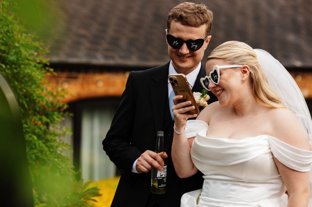Happy bride and groom in wedding attire enjoying a moment outdoors, smiling and looking at a smartphone, with a rustic building in the background.