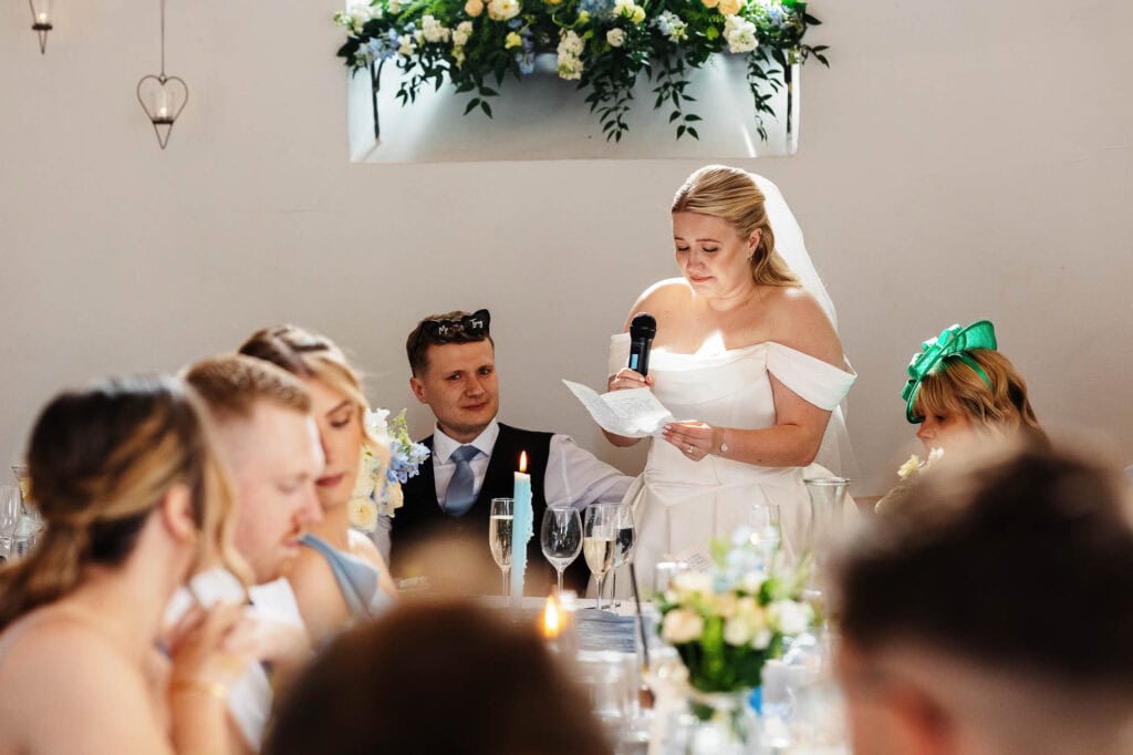 A1: Elegant bride giving a speech at her wedding reception surrounded by friends and family, with floral decorations and candles creating a warm, celebratory atmosphere.