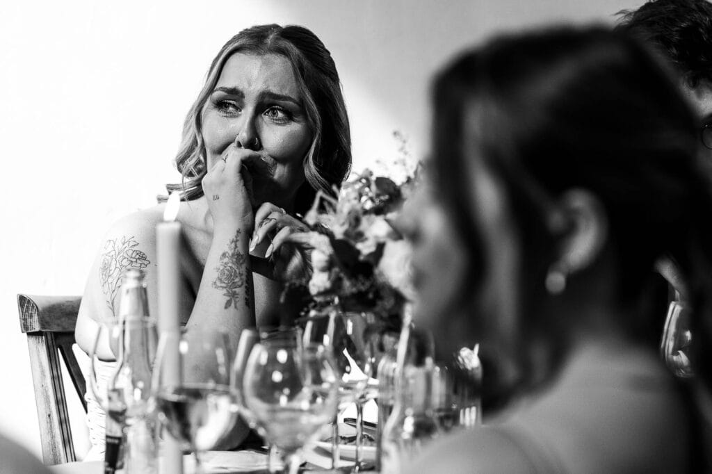 A1. Emotional bride at a wedding reception, expressing joy and tears, surrounded by wine glasses and friends, captured in black and white for a timeless wedding moment.