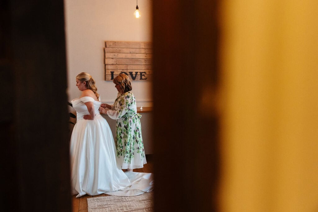 Elegant bride in a white wedding gown with a floral guest preparing her at a wedding venue, capturing a special moment before the ceremony.