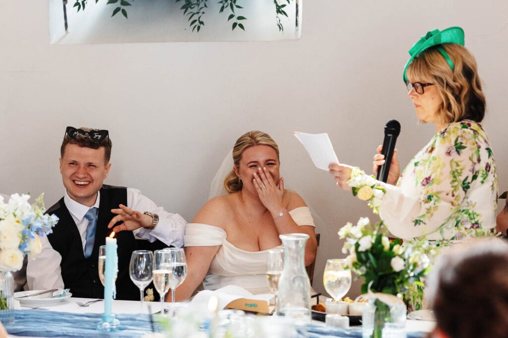 Joyful wedding reception with bride, groom, and guest giving speech, decorated with flowers and elegant table settings, capturing a happy moment in a bright, modern venue.