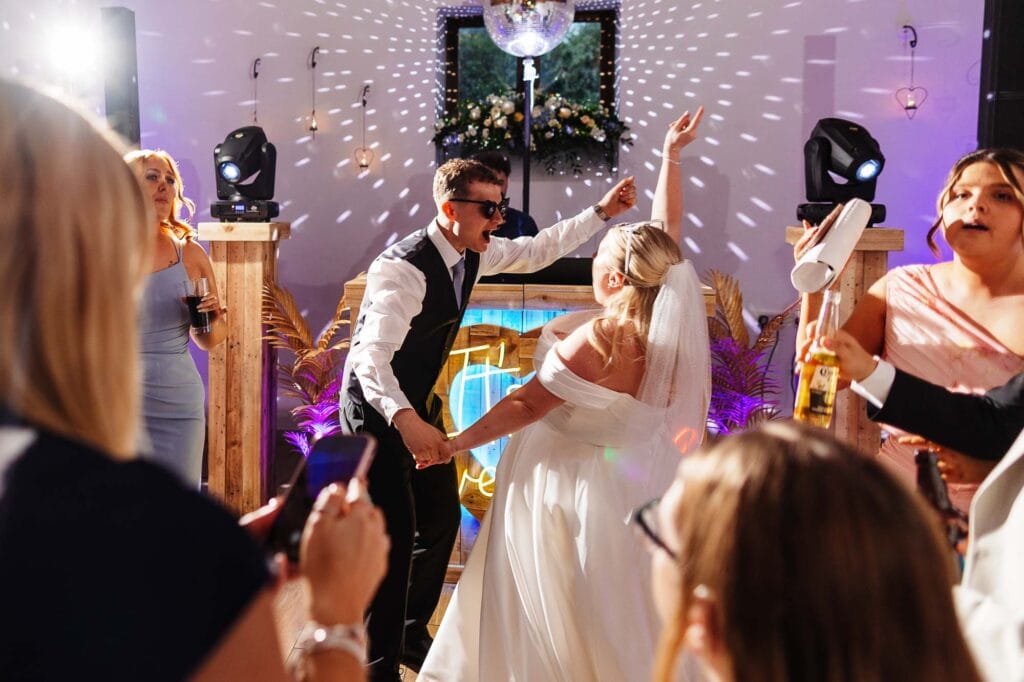 Joyful wedding reception with dancing bride and groom, surrounded by friends, colourful lighting, and lively atmosphere at BGS Weddings in the UK.