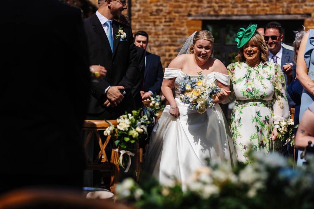 A1. Elegant bride in a white wedding gown holding a bouquet, surrounded by joyful guests at an outdoor wedding ceremony in a historic venue.