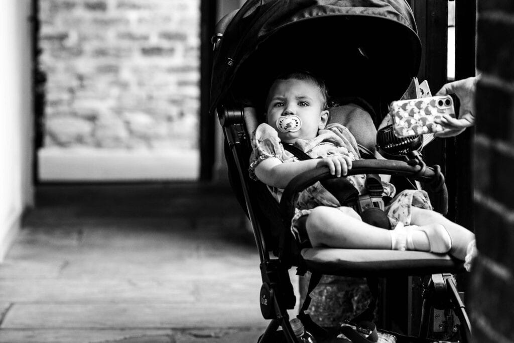 Black and white photo of a young girl sitting in a stroller with a pacifier, holding a gift box, at a wedding reception or outdoor event in the UK.
