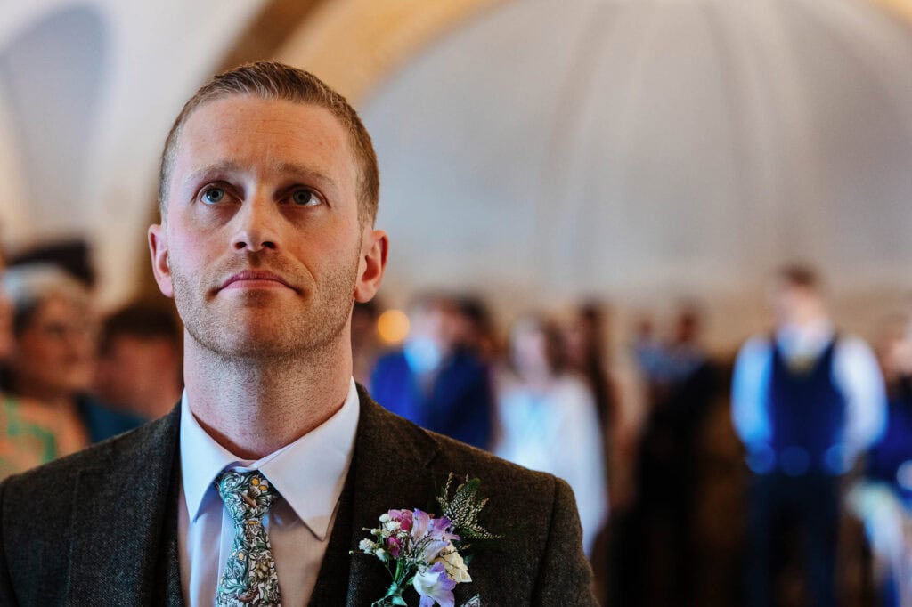 Close-up of a groom during a wedding ceremony, wearing a dark suit with a floral boutonniere, in a beautifully decorated venue with guests in the background.