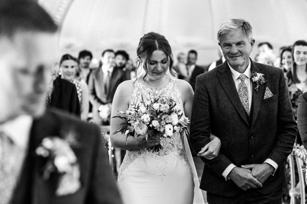 Beautiful black and white image of a bride walking down the aisle with her father at a wedding ceremony, surrounded by guests, captured at BGS Weddings.