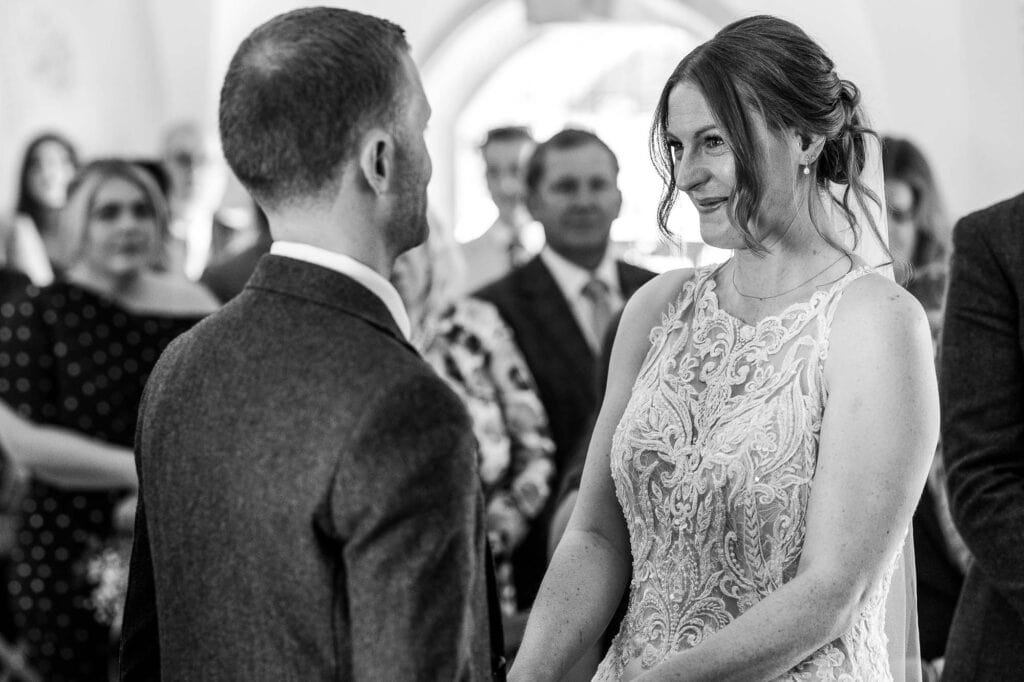 Bride and groom sharing vows during their wedding ceremony, surrounded by family and friends, capturing a heartfelt moment in black and white.