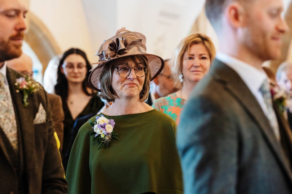 A woman wearing a stylish hat and glasses attending a wedding ceremony, surrounded by guests in formal attire, capturing a memorable moment of the special day.