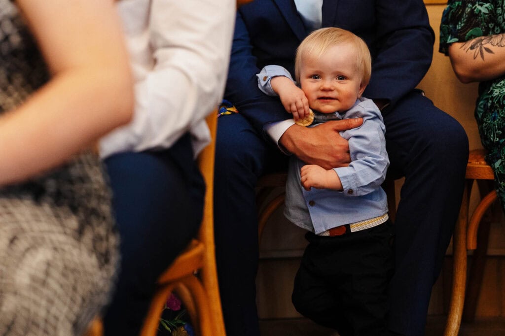 Young boy smiling and holding onto an adult's hand during a wedding ceremony, surrounded by seated guests, capturing a candid moment of childhood innocence and happiness.