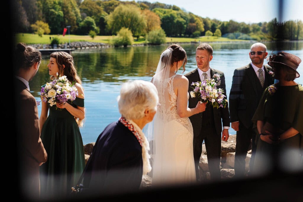 Wedding ceremony by the river with bride, groom, and guests at BGS Weddings, showcasing a scenic outdoor setting with lush greenery and calm water, perfect for romantic wedding celebrations.
