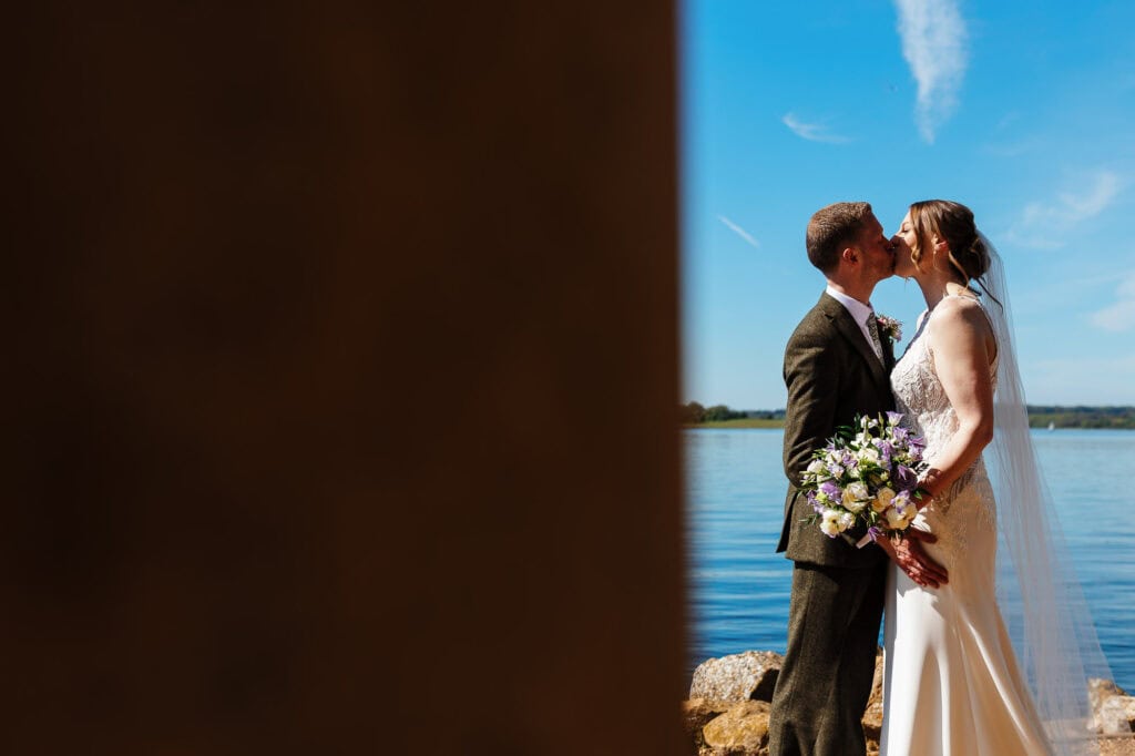 Romantic wedding couple sharing a kiss by the lake with a scenic water backdrop and blue sky, capturing a special moment on their wedding day.