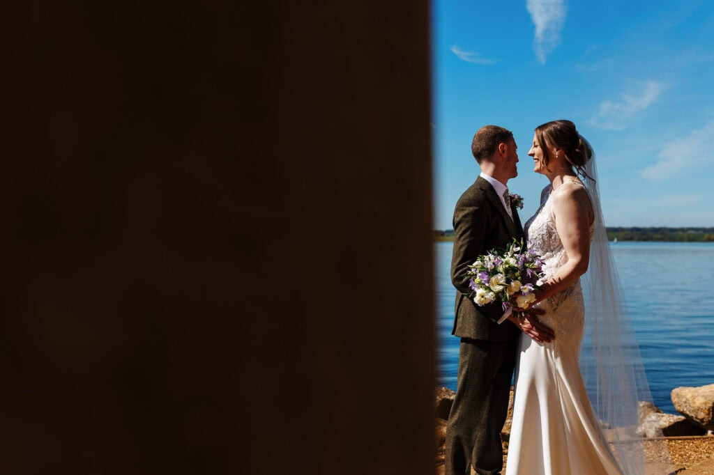 Romantic wedding couple standing by a lake, with the bride holding a bouquet and the groom in a suit, during a beautiful outdoor ceremony at BGS Weddings.