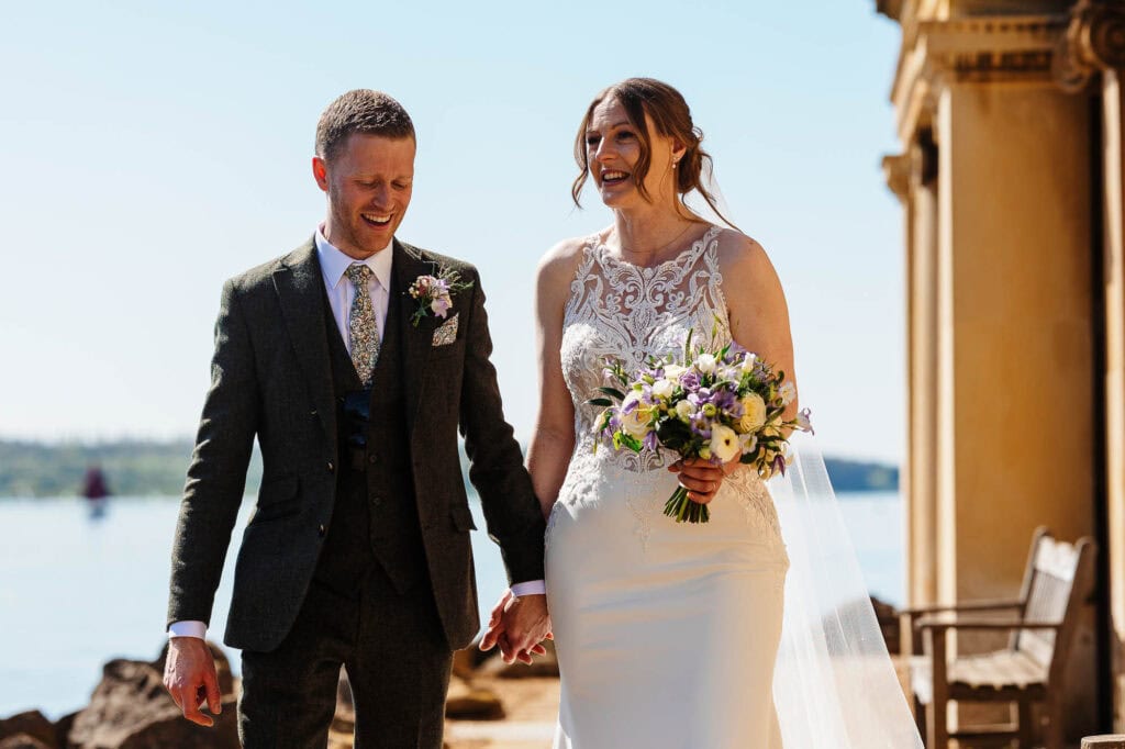 A joyful bride and groom holding hands by the waterfront during their wedding at BGS Weddings, showcasing a beautiful outdoor setting with elegant attire and a scenic background.