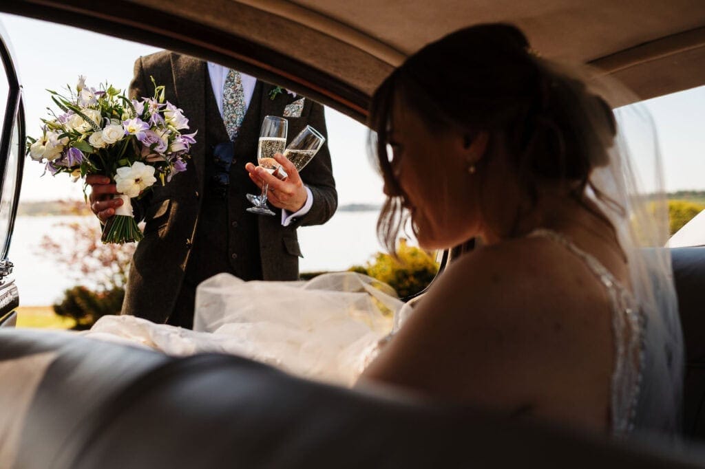 Wedding couple toasting with champagne, holding a bouquet, with scenic outdoor background.