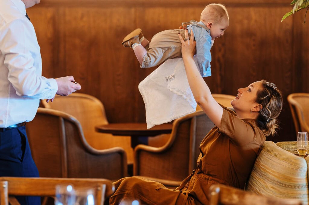 Woman lifting a smiling baby in a restaurant setting with wooden decor and dining tables, capturing a joyful family interaction during a wedding celebration.