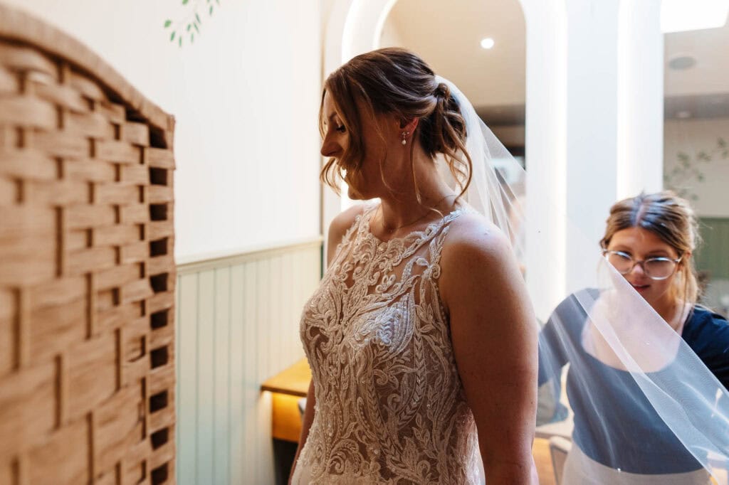 Bride in lace wedding gown during fitting session, with assistant helping her in a bright, elegant bridal boutique.
