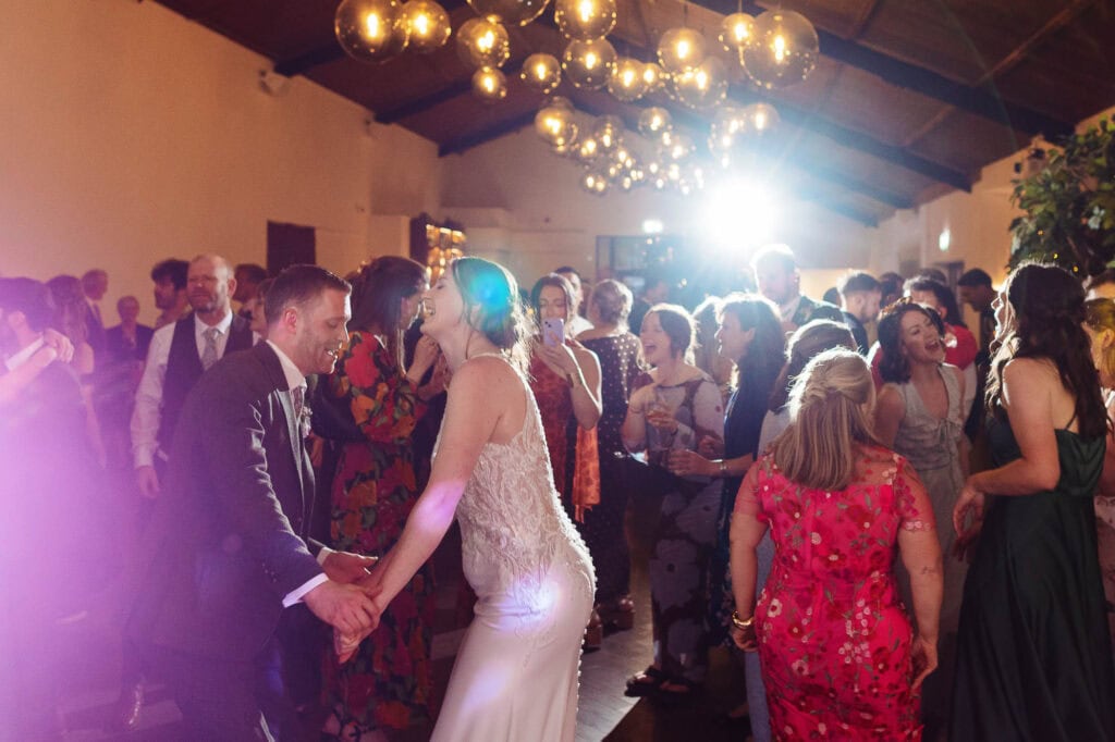 A lively wedding celebration featuring a bride and groom dancing surrounded by friends and family in a beautifully decorated venue. The atmosphere is joyful, with vibrant lighting and elegant decor.
