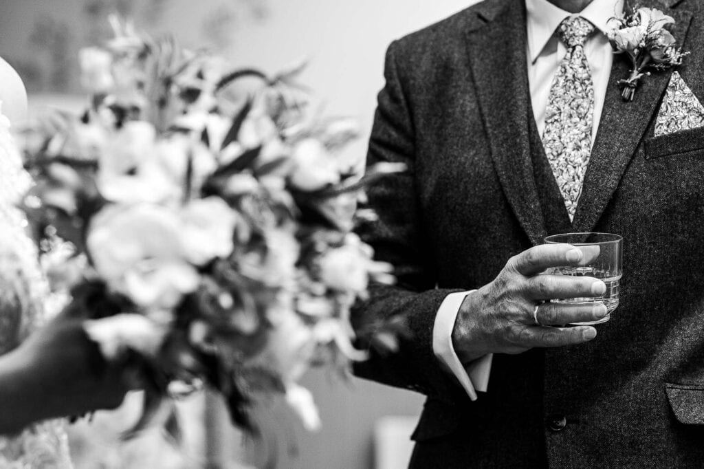 Close-up of groom holding a glass during wedding toast, with floral bouquet in foreground, in black and white.