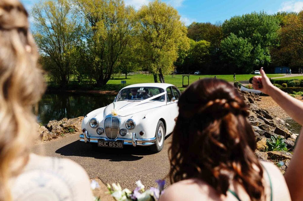 Elegant white wedding car parked near a pond with lush green trees in the background.