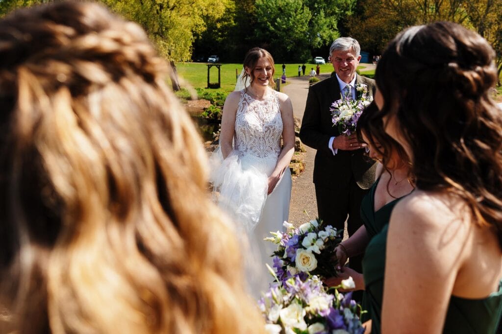 Bride and groom with wedding party outdoors in a lush green park, smiling and holding flowers during a sunny wedding ceremony.