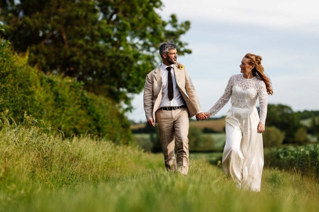 Beautiful wedding couple walking hand in hand through a lush green field on their special day, surrounded by nature and clear skies.