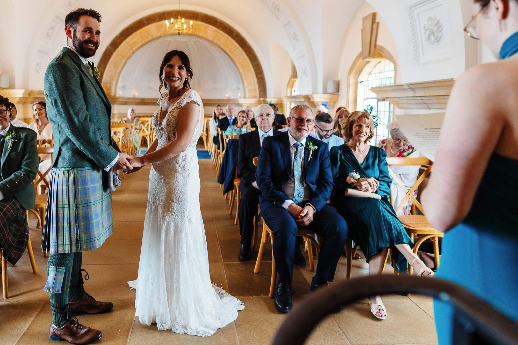 Beautiful bride and groom exchanging vows during a wedding ceremony in an elegant, historic venue with guests seated and witnessing the special moment.