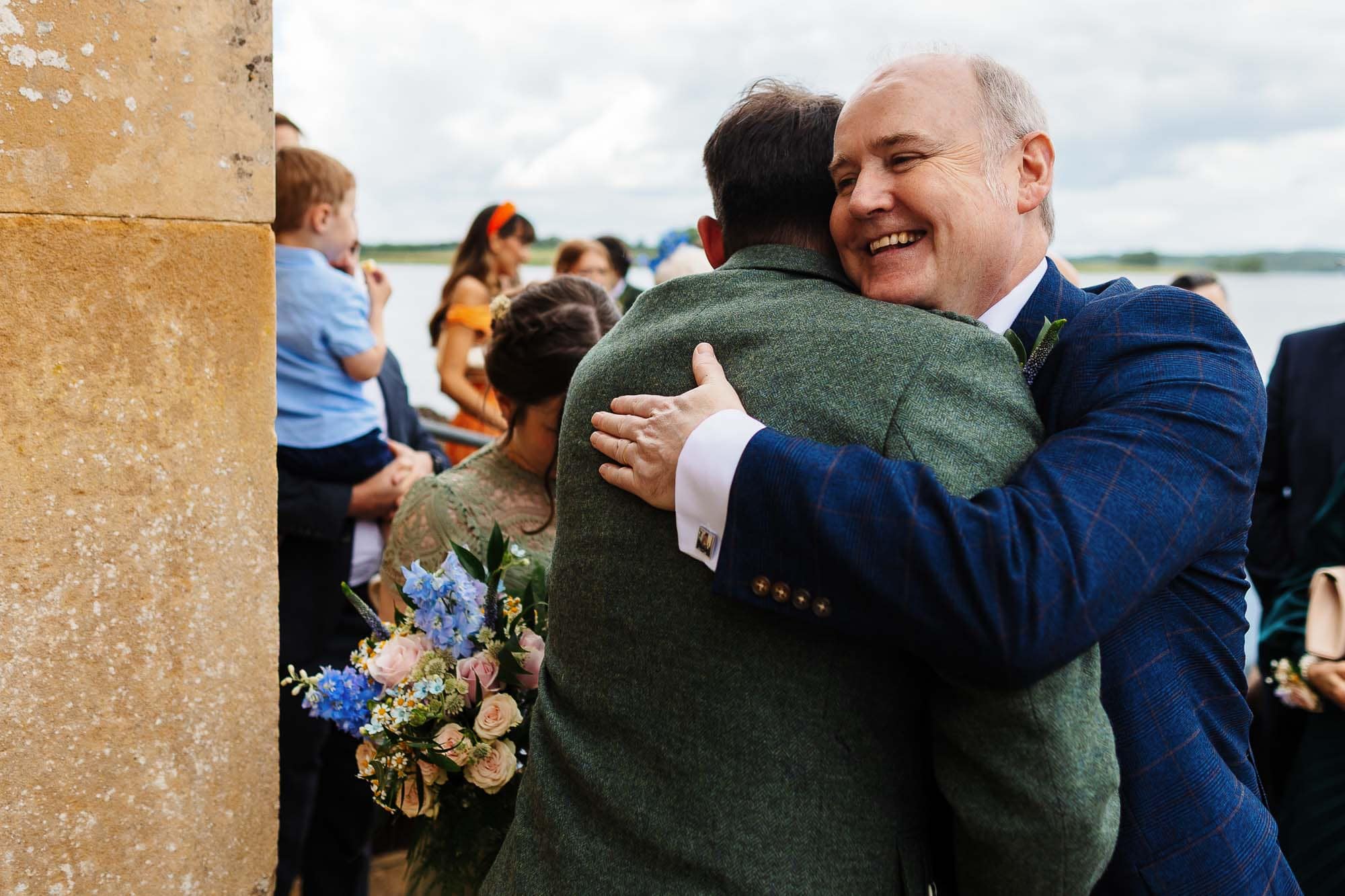 Happy groom embracing a guest during a wedding celebration outdoors, with friends and family in the background, capturing joyful moments at BGS Weddings.